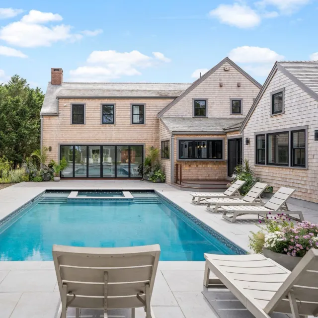 Modern house with multiple windows and wood siding, backyard with swimming pool, poolside chairs, and potted plants, under a partly cloudy sky.