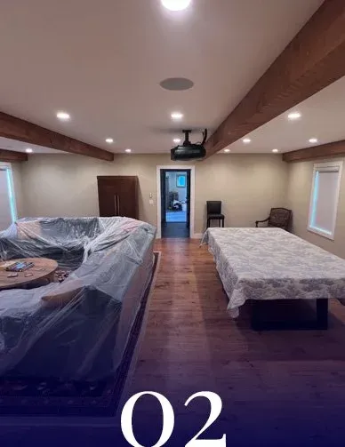 A bedroom with a sloped ceiling, two beds (one with plastic covering), hardwood floors, and two small windows. There is a door in the center leading to another room and a ceiling-mounted projector. Simple furniture includes a chair and a nightstand.