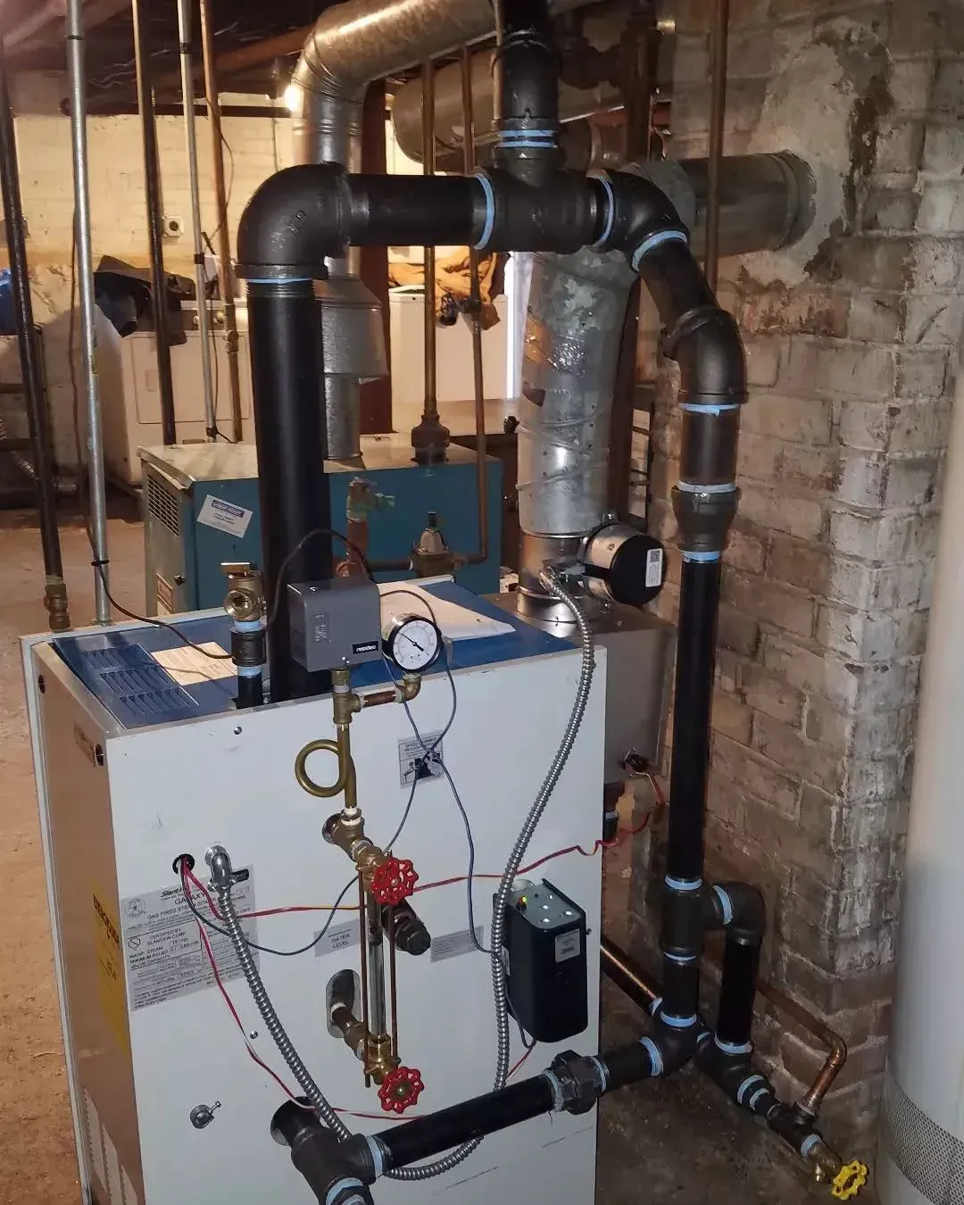 Boiler room machinery with white control panel, gauges, and black pipes against a brick wall.