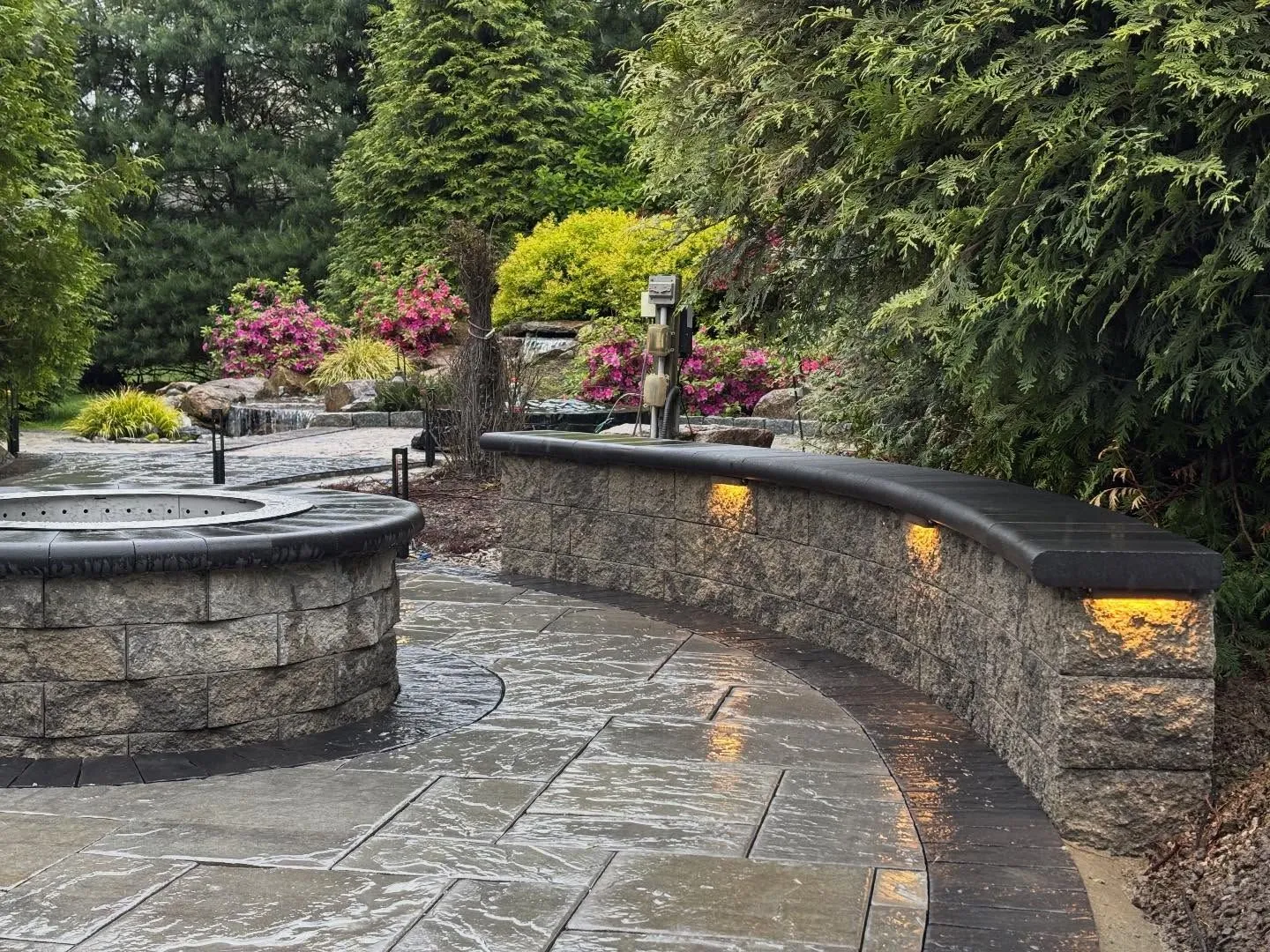 Curved stone garden benches beside a wet paved path in a lush landscaped park with flowers and trees