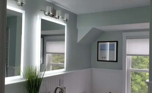 A bathroom with a large backlit mirror, two windows with white blinds, a framed beach scene picture, a green plant, and a three-light fixture above the mirror.