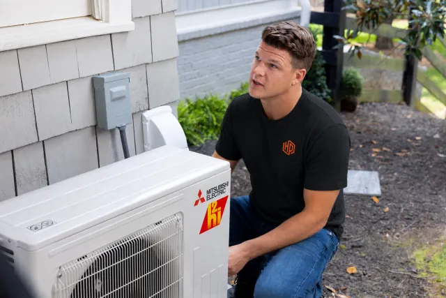 A man wearing a black t-shirt with a yellow and orange logo kneeling outside near an air conditioning unit with Mitsubishi Electric and hi stickers, on the side of a house with gray siding, doing maintenance or inspection.