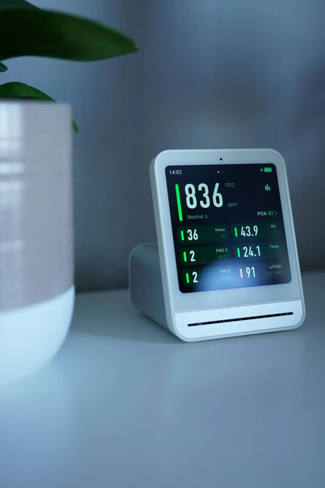 Digital bedside clock on a gray table next to a white plant pot, showing 8:36 AM and weather data.
