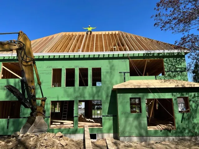 A person in a yellow safety vest sits atop the roof rafters of a house under construction with an excavator in the front.