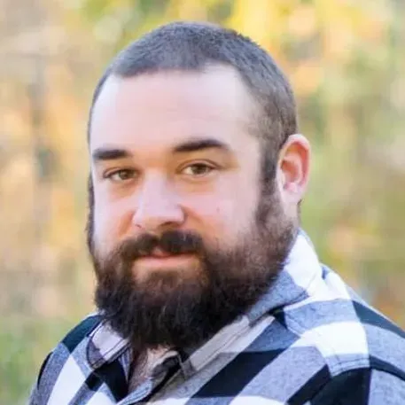 Man with short dark hair and full beard wearing a black and white checkered shirt outdoors.