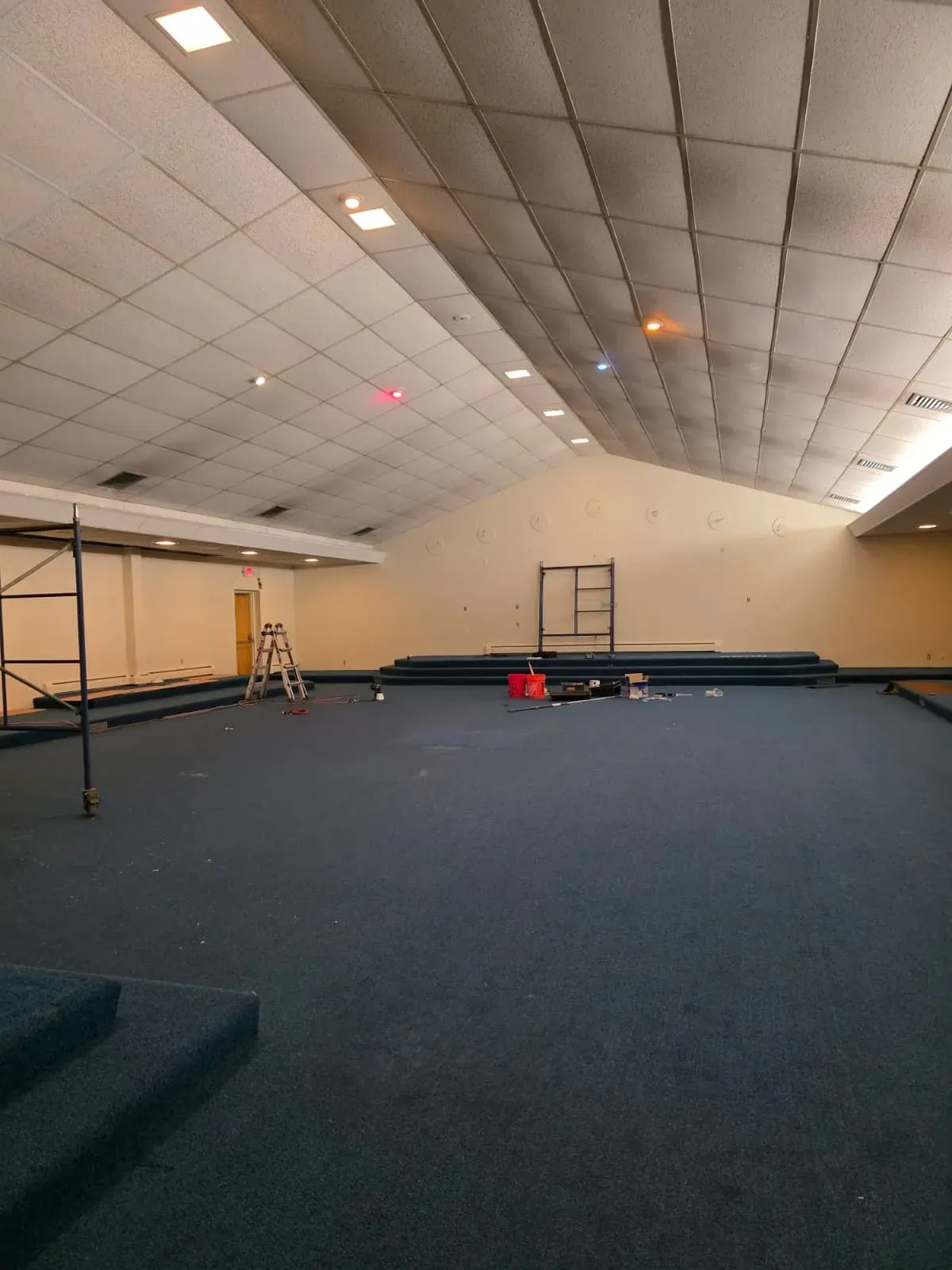 Large empty indoor hall with blue carpet, sloped ceiling, and a few ladders and equipment near the back wall