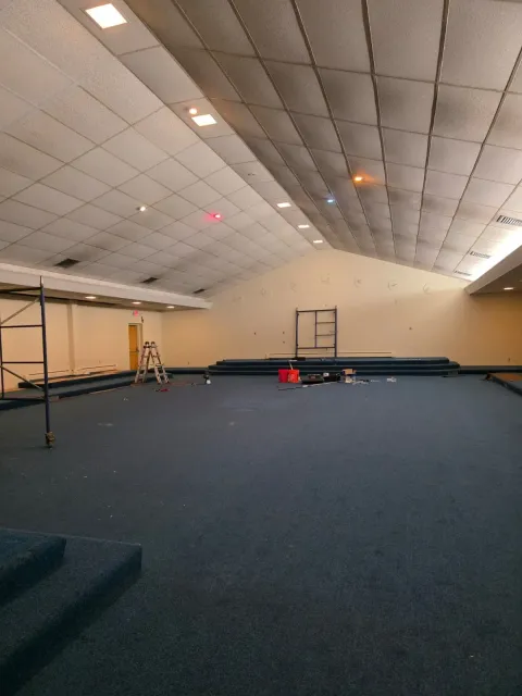 Large empty indoor hall with blue carpet, sloped ceiling, and a few ladders and equipment near the back wall
