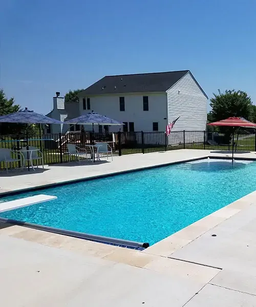 A clear blue swimming pool with white deck chairs, umbrellas, and tables around it, set in a fenced backyard with a two-story white house in the background under a clear blue sky.
