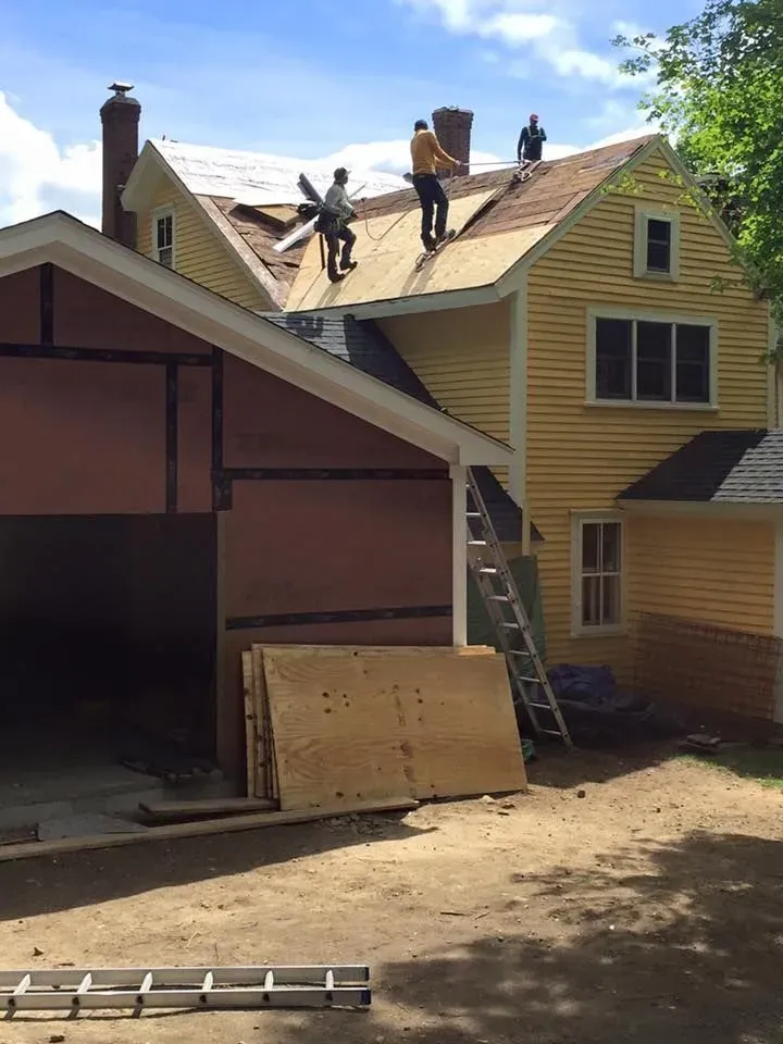 Roofers on a yellow house, removing old shingles. A ladder leans against the building; construction in progress.