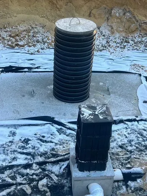 Black cylindrical vent stack and a smaller wrapped pipe on concrete in a snowy outdoor area.