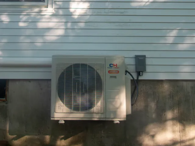 Exterior of a house with an air conditioning unit mounted on the wall near the ground, with a weathered concrete surface below and white horizontal siding on the house wall.