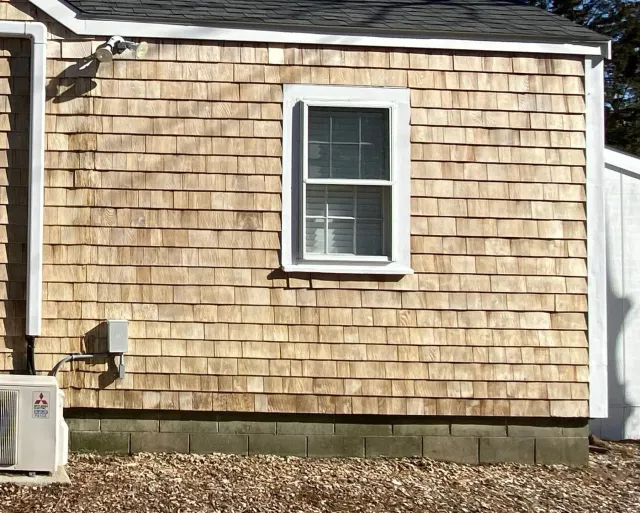Wood-shingled building with white-framed window, air conditioner, and electrical box; gravel ground.