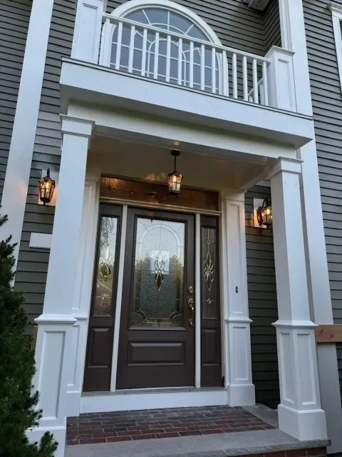 Front porch of a house with a dark wooden door featuring decorative glass panels, white framing, and exterior lighting fixtures on either side of the entrance. There is a balcony with a white railing above.