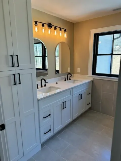 A bathroom with white cabinets, double vanity sinks, arched mirrors, a five-light fixture, and a black-framed window.
