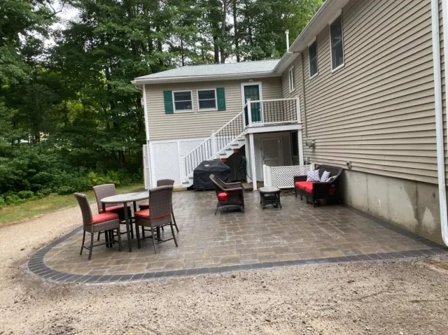 A beige house with a stone patio, outdoor dining set, and seating area in a backyard surrounded by woods.