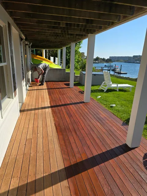 Wooden deck by the water with white chairs, a kayak, and a person near the shaded patio.