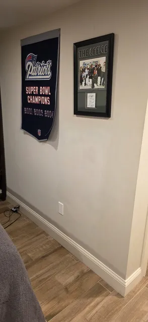 View of a finished interior wall with white baseboard trim and wood-look floor tiles. Features a Patriots banner and framed Office poster. Handyman Boston services for home finishing and details.