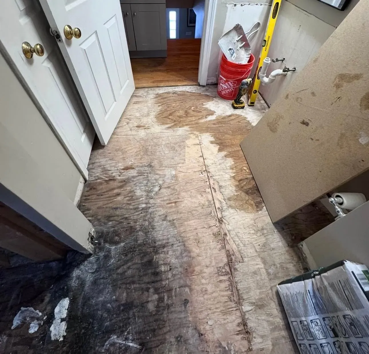 Water-damaged hallway floor with dark stains and peeling surface, beside an open door and scattered debris