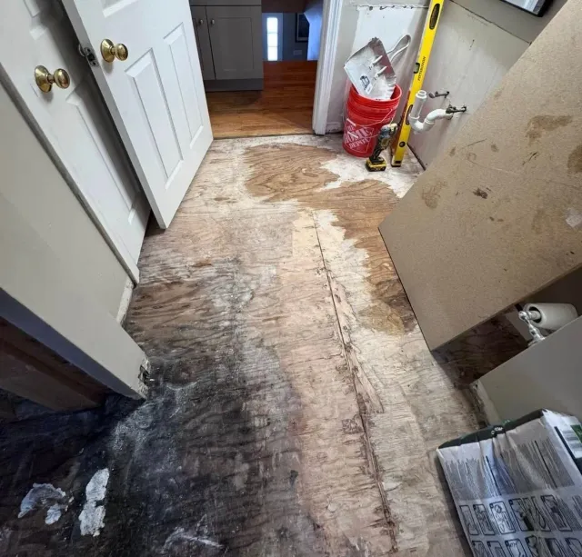 Water-damaged hallway floor with dark stains and peeling surface, beside an open door and scattered debris