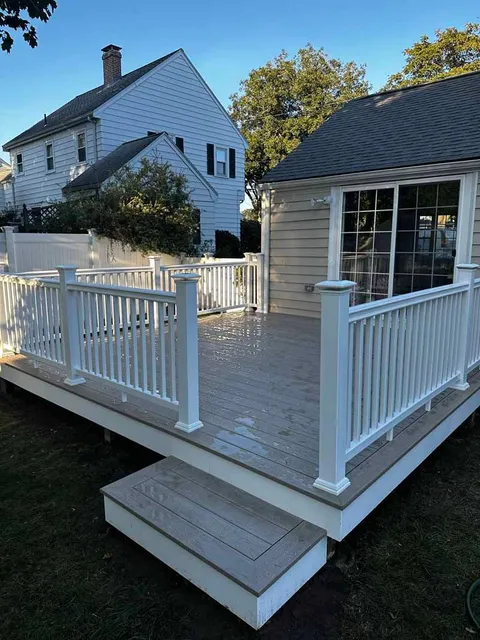 White-railed wooden deck with stairs in a yard, next to a beige building, with a house in the background.