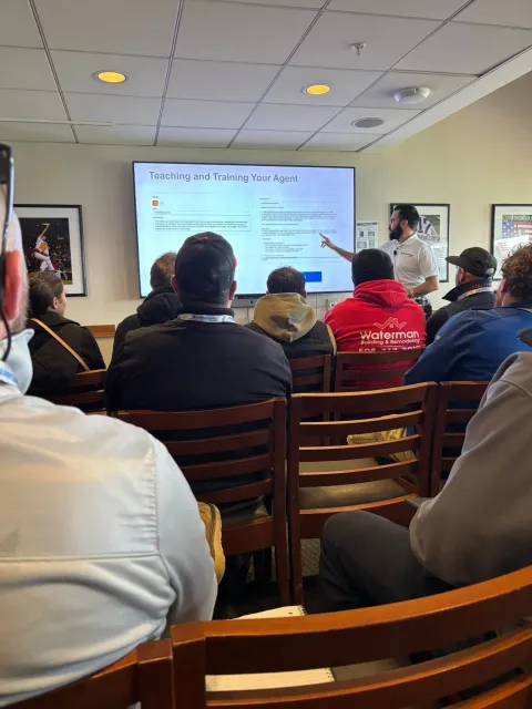 People seated in a meeting room watching a presentation on a projector screen.