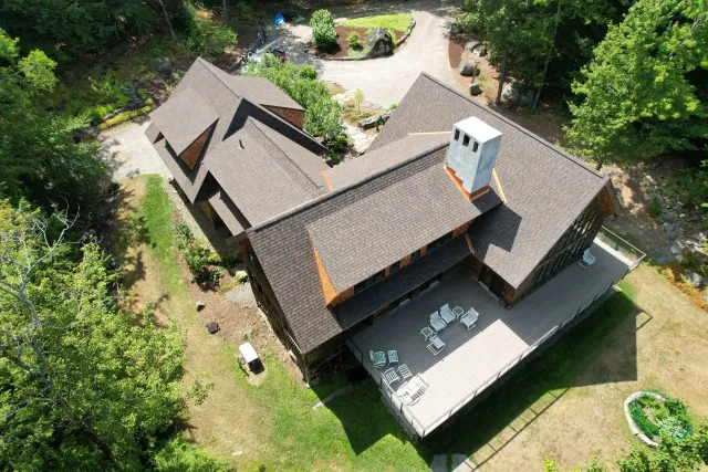 Aerial view of a brown-shingled house with a deck, surrounded by trees and lawn.