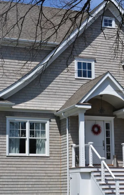 The front of a home with gray shingle siding and a steep gable roof with dormer windows and a small porch.