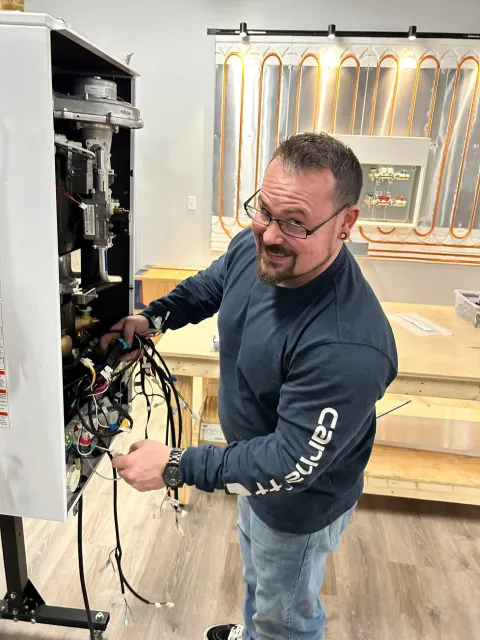 Man working inside an open electrical panel in a workshop, smiling at the camera