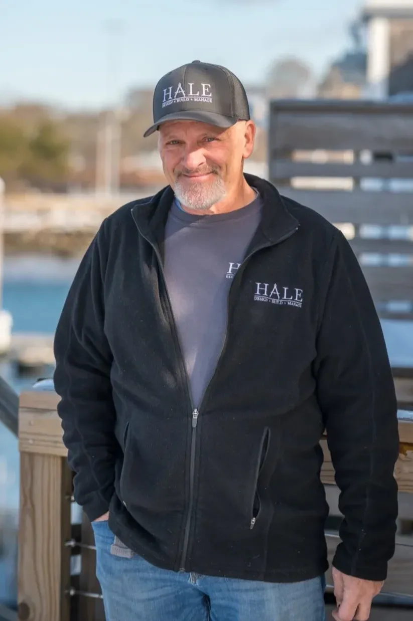 A smiling man with a beard, wearing a black Hale cap and jacket, and a gray shirt, standing outdoors on a dock with a body of water and blurred background behind him.