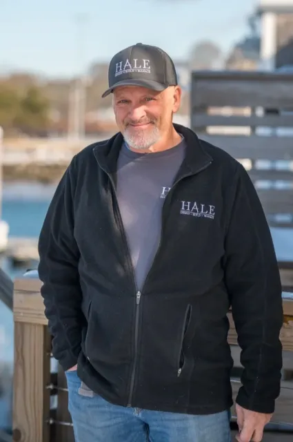 A smiling man with a beard, wearing a black Hale cap and jacket, and a gray shirt, standing outdoors on a dock with a body of water and blurred background behind him.