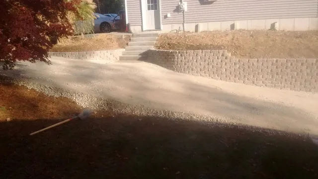 A completed residential landscaping project in Dudley featuring a curved tan masonry retaining wall, a gravel driveway, and concrete stairs leading to a house with light-colored siding. A Japanese Maple tree with red leaves adds vibrant color to the scene.