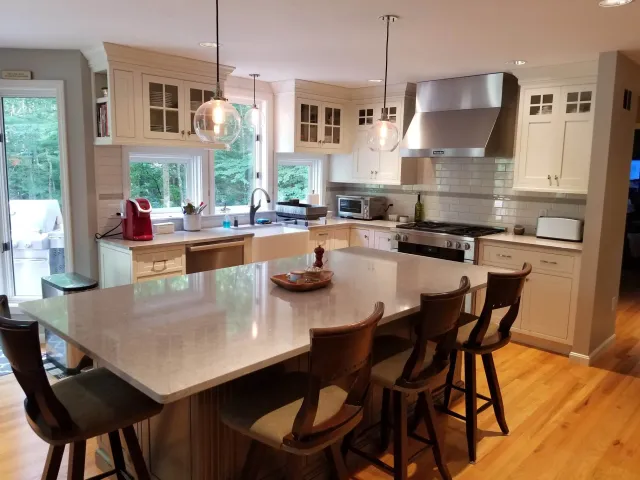 Kitchen with a large central island, four wooden bar stools, white cabinets, stainless steel range hood, stainless steel appliances, pendant lights, and large windows showing a wooded backyard.