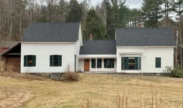 White farmhouse with dark shutters and roof, brown door, set in a field.