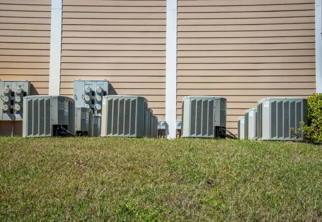 Air conditioning units on a grassy area in front of a light brown textured wall.