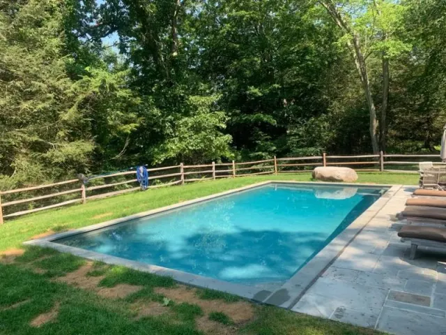 A rectangular backyard swimming pool with blue water, surrounded by a stone patio, green grass, and a wooden fence.