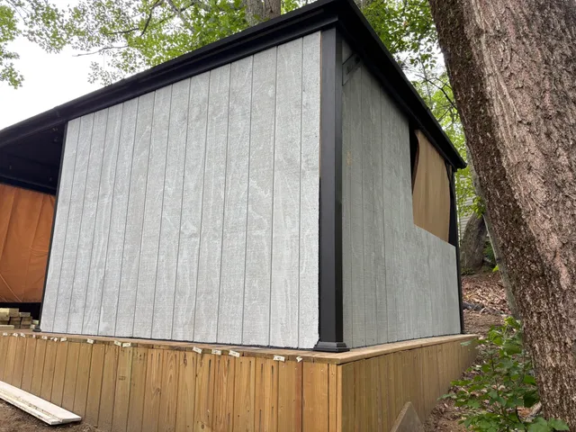 Screened porch framing in-progress build with gray siding, black trim, and wooden foundation, surrounded by natural foliage.
