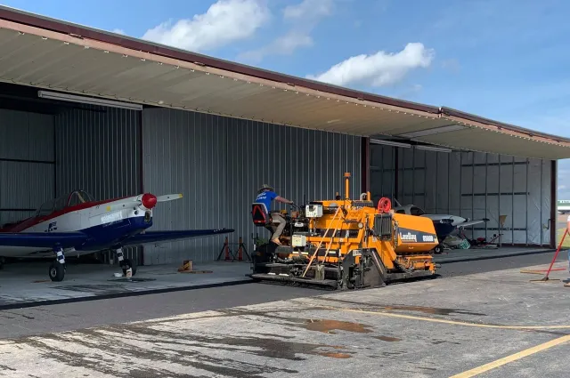 A light airplane and a large yellow construction vehicle parked inside an open metal hangar.
