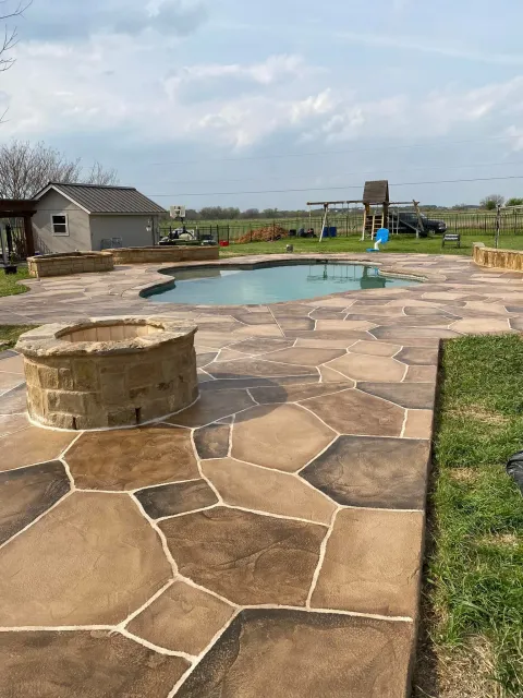 Stone patio with fire pit, pool, small building, and play set under cloudy sky.