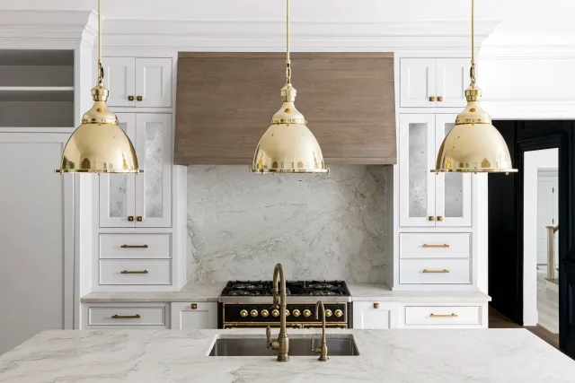 Modern kitchen with white cabinetry, marble backsplash and countertops, brass pendant lights, and a black and gold stove.