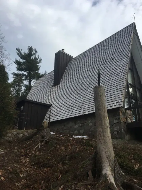 A-frame house with stone base and cedar shake roof, set on a hillside with a dead tree in the foreground.