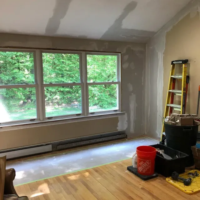 Partially renovated room with large windows, patchy drywall, ladder, tools, and a bucket on the floor