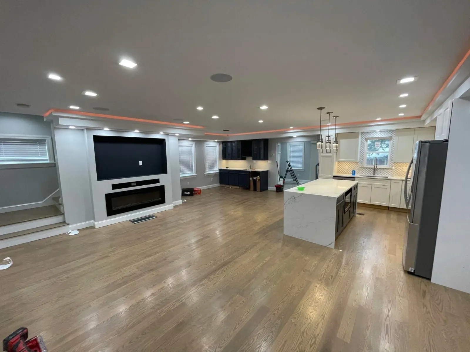 Open-concept basement with hardwood floors, a large central island, a kitchen with white cabinets, a stainless steel refrigerator, and a wall-mounted TV above an electric fireplace, with ongoing construction.