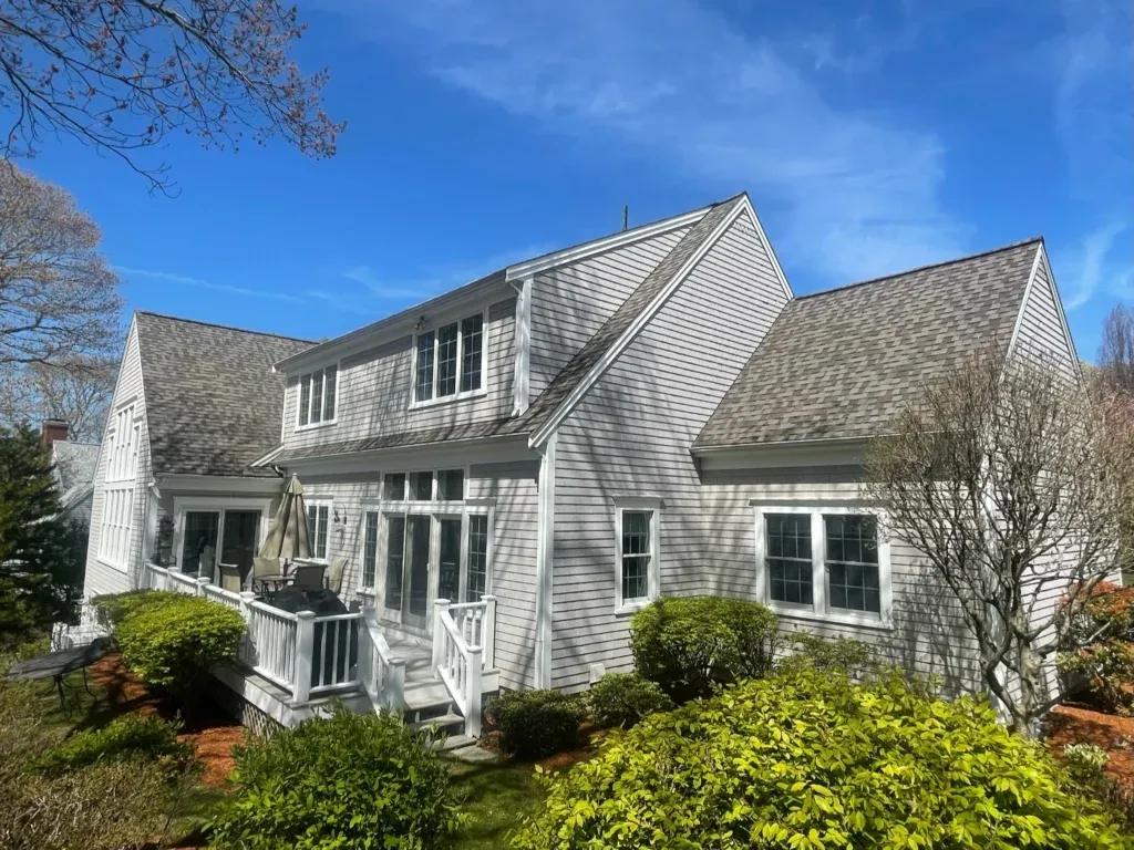 A spacious white house with gabled roofs and multiple large windows. A small patio with a table and chairs is visible, surrounded by lush green shrubs and trees. The sky is clear and bright blue, indicating a sunny day.