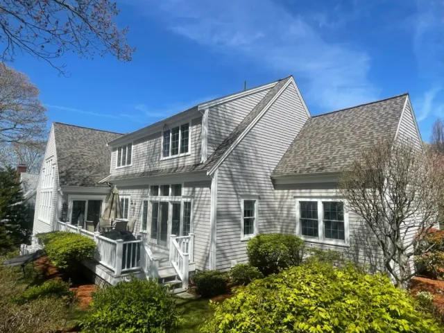 A spacious white house with gabled roofs and multiple large windows. A small patio with a table and chairs is visible, surrounded by lush green shrubs and trees. The sky is clear and bright blue, indicating a sunny day.