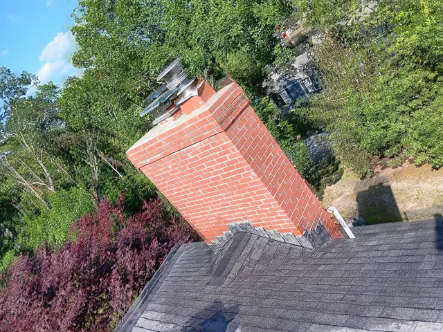 Leaning brick chimney on a roof with a dark shingled surface, surrounded by green trees and bushes.