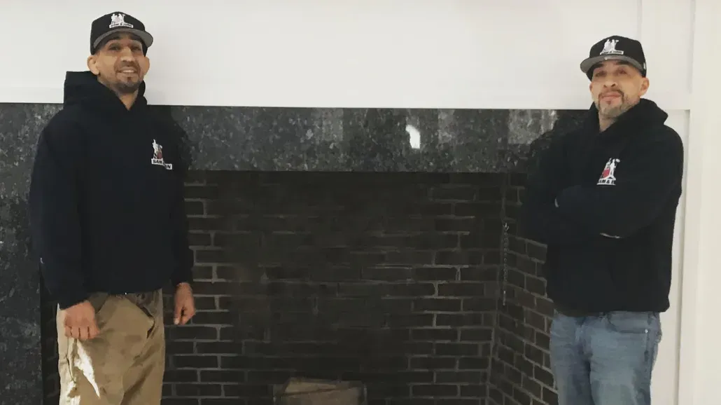 Two men in black jackets and caps standing beside a dark brick fireplace mantle