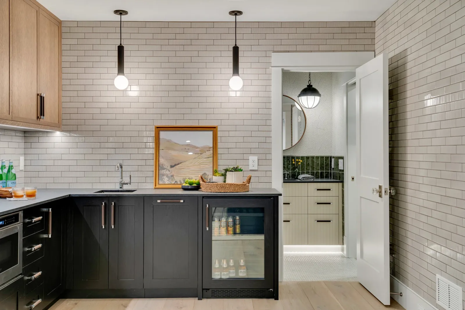 Hardwood floor installation in a Norwell kitchen with a beige brick backsplash, black lower cabinets, pendant lighting, a framed landscape painting, and a view into a bathroom with mirrored black and green tile.