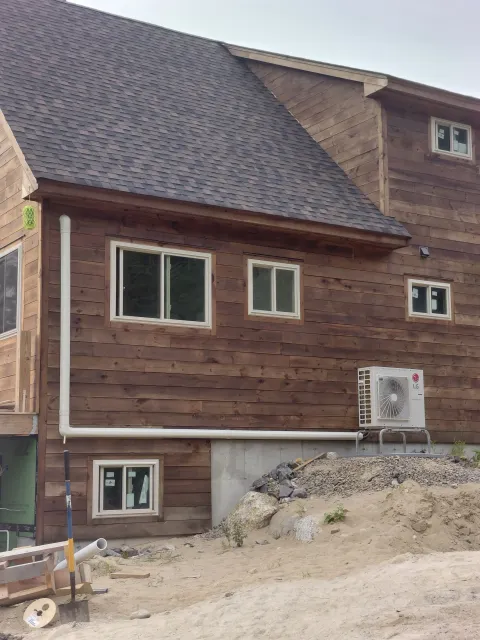 A house under construction with wooden siding, multiple small windows, and an air conditioning unit outside. There is construction debris and dirt in the foreground.