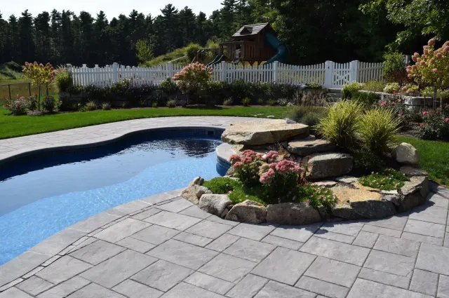 A landscaped backyard with a pool, stone patio, rock steps, and white picket fence.