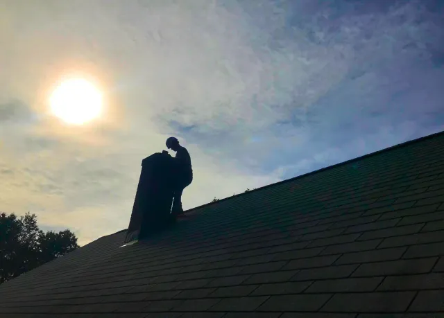 Silhouetted worker in a helmet standing on a roof near a chimney, with the sun setting or rising in the background and a partly cloudy sky.